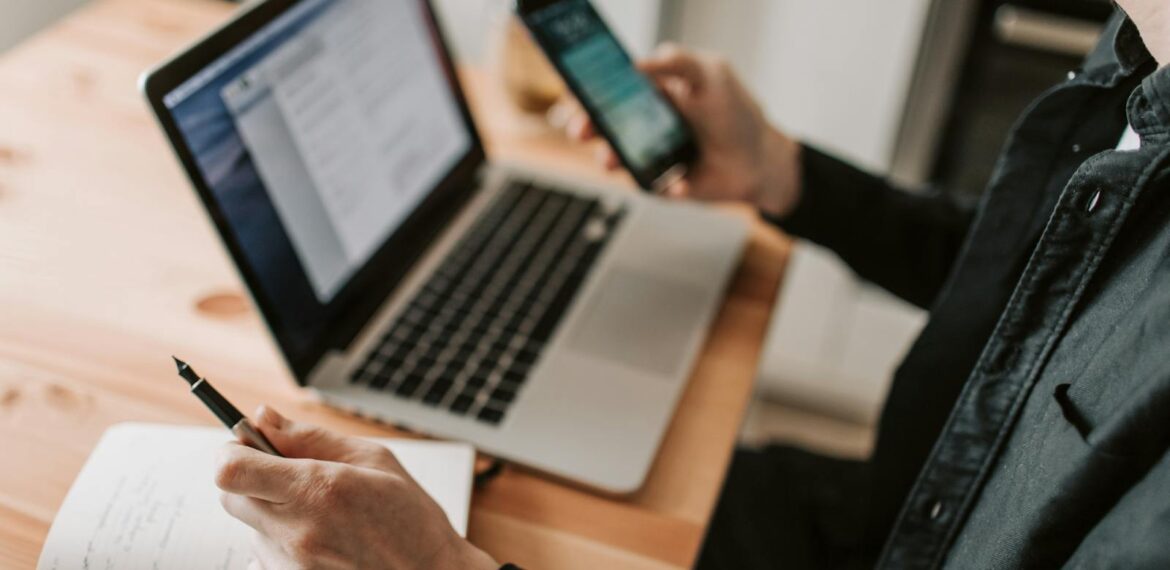 A person multitasking with a laptop and smartphone at a wooden desk.